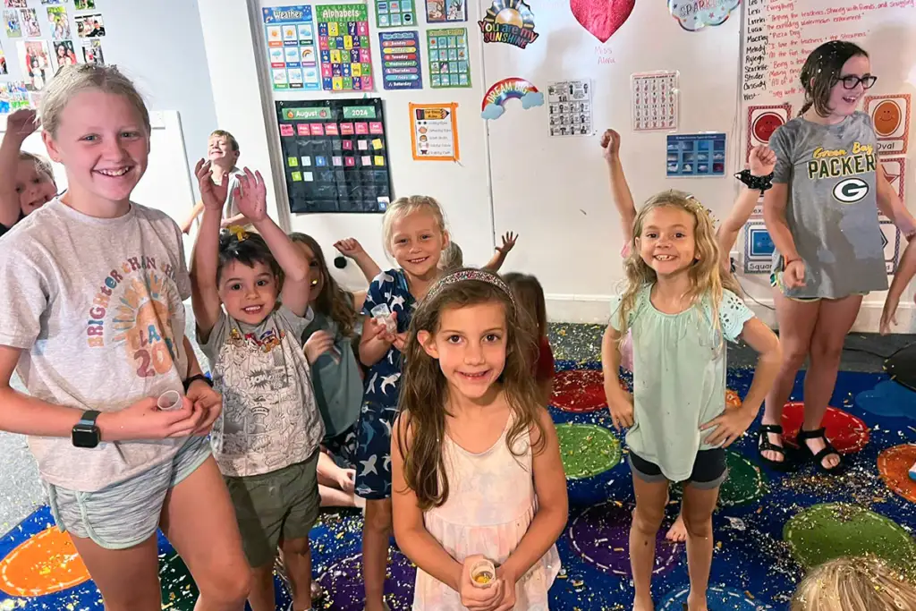 A group of smiling children in a colorful classroom celebrating with glitter and confetti.